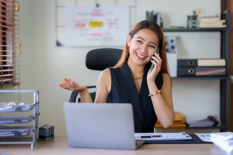 Mulher sorridente falando ao celular enquanto trabalha, contente porque está evitando a inadimplência de clientes.