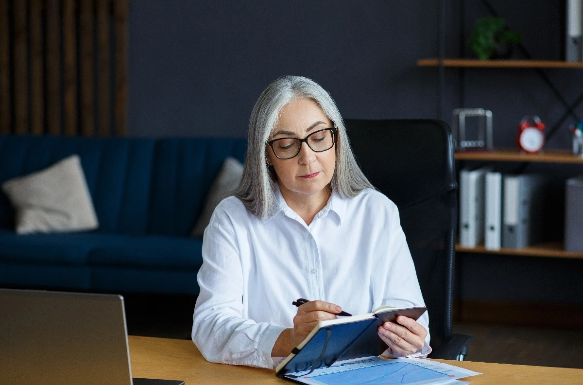 Mulher sorridente, com cabelos grisalhos, em home office, escrevendo e estudando se mei tem direito a aposentadoria.